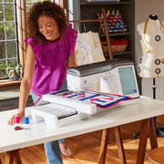 A smiling creator working with colorful fabrics at a bright craft desk alongside the Brother Stellaire Innov-is XJ2 machine