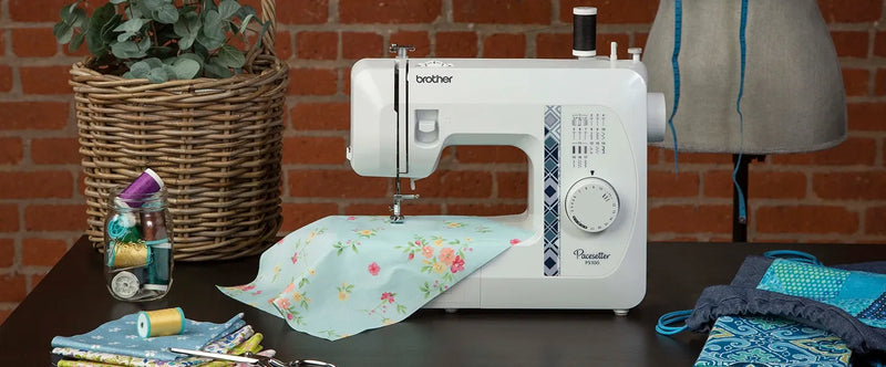 brother-pacesetter-ps100-sewing-machine-with-floral-light-blue-fabric-on-table-next-to-woven-basket-and-thread-jar
