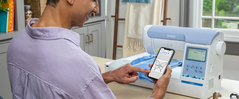 man-holding-smartphone-with-artspira-app-displaying-butterfly-pattern-next-to-brother-pe900-embroidery-machine