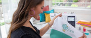 smiling-woman-holding-white-and-green-tote-bag-with-custom-cactus-and-ava-name-embroidery-next-to-brother-se700-machine