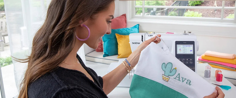 smiling-woman-holding-white-and-green-tote-bag-with-custom-cactus-and-ava-name-embroidery-next-to-brother-se700-machine