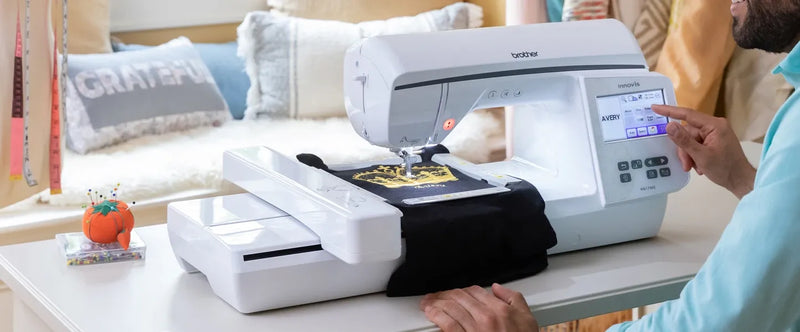 side-angle-view-user-finger-navigating-menu-on-brother-innov-is-nq1700e-embroidery-machine-stitching-gold-crest-design-on-black-fabric